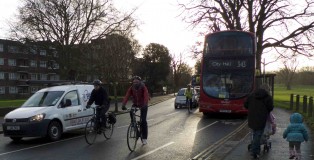 Cyclists with motor cars and bus in cycle path. Person pushes pram with toddler next to them