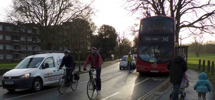 Cyclists with motor cars and bus in cycle path. Person pushes pram with toddler next to them
